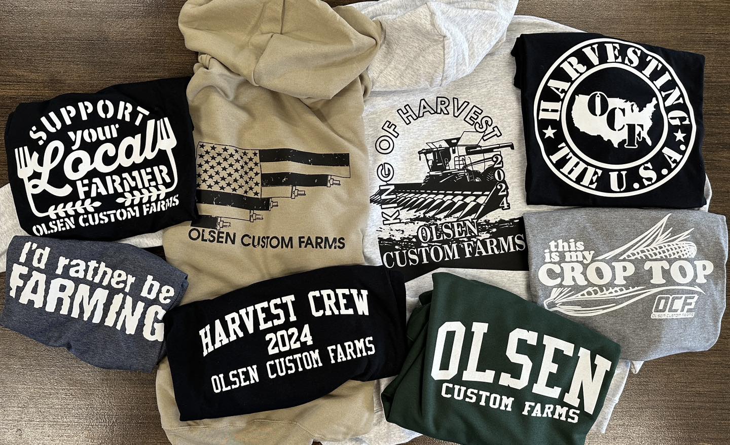 Collection of screen-printed t-shirts and hoodies with 'Olsen Custom Farms' branding on a wooden surface.