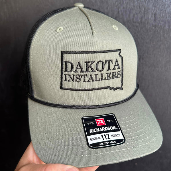 Gray cap with 'Dakota Installers' logo held against a dark background made in Volga SD