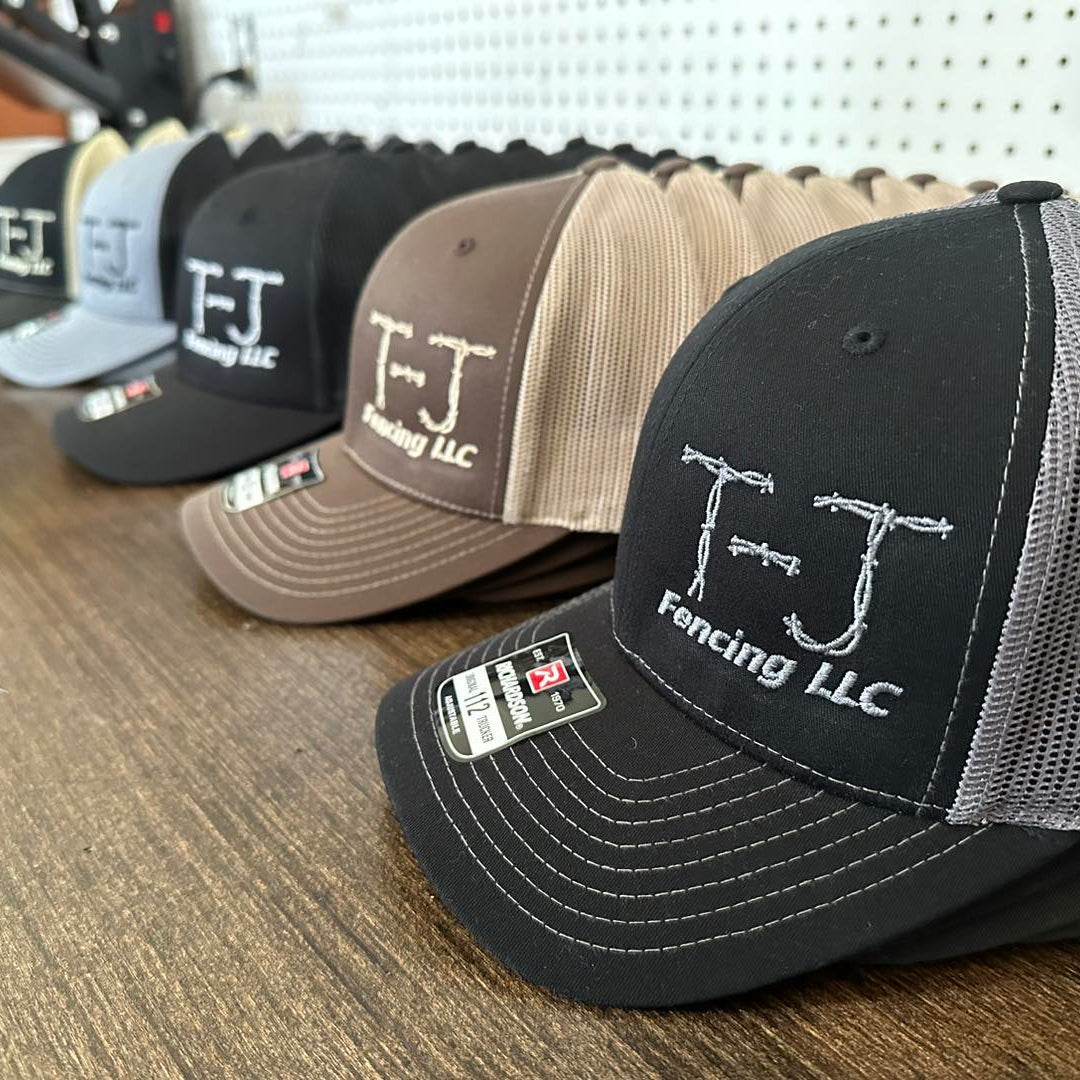 Row of embroidered TJ Fencing LLC caps on a wooden surface in Volga, SD