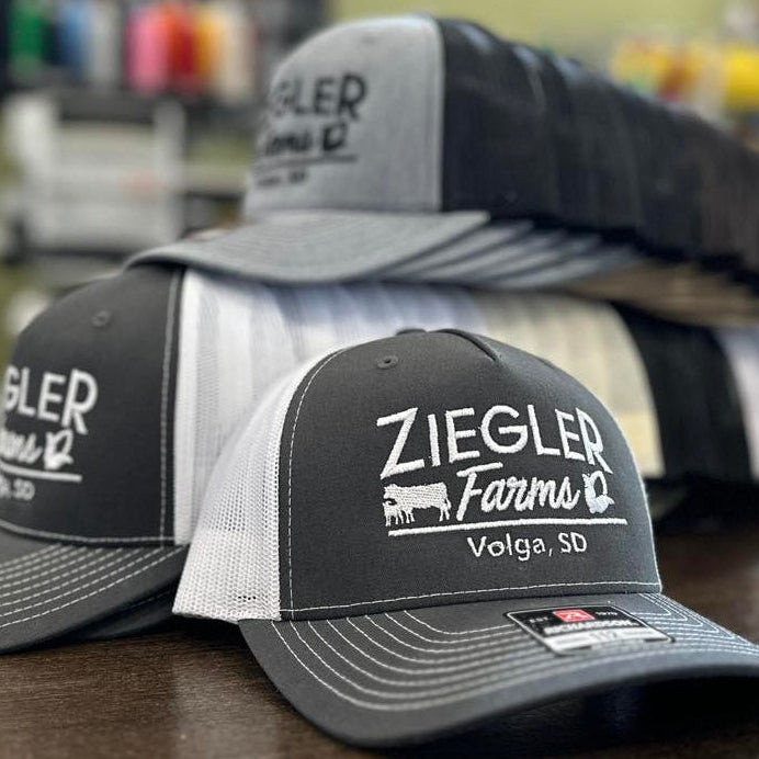 Gray and black Richardson caps with 'Ziegler Farms Volga, SD' logo on a wooden surface made by Cotton & Customs