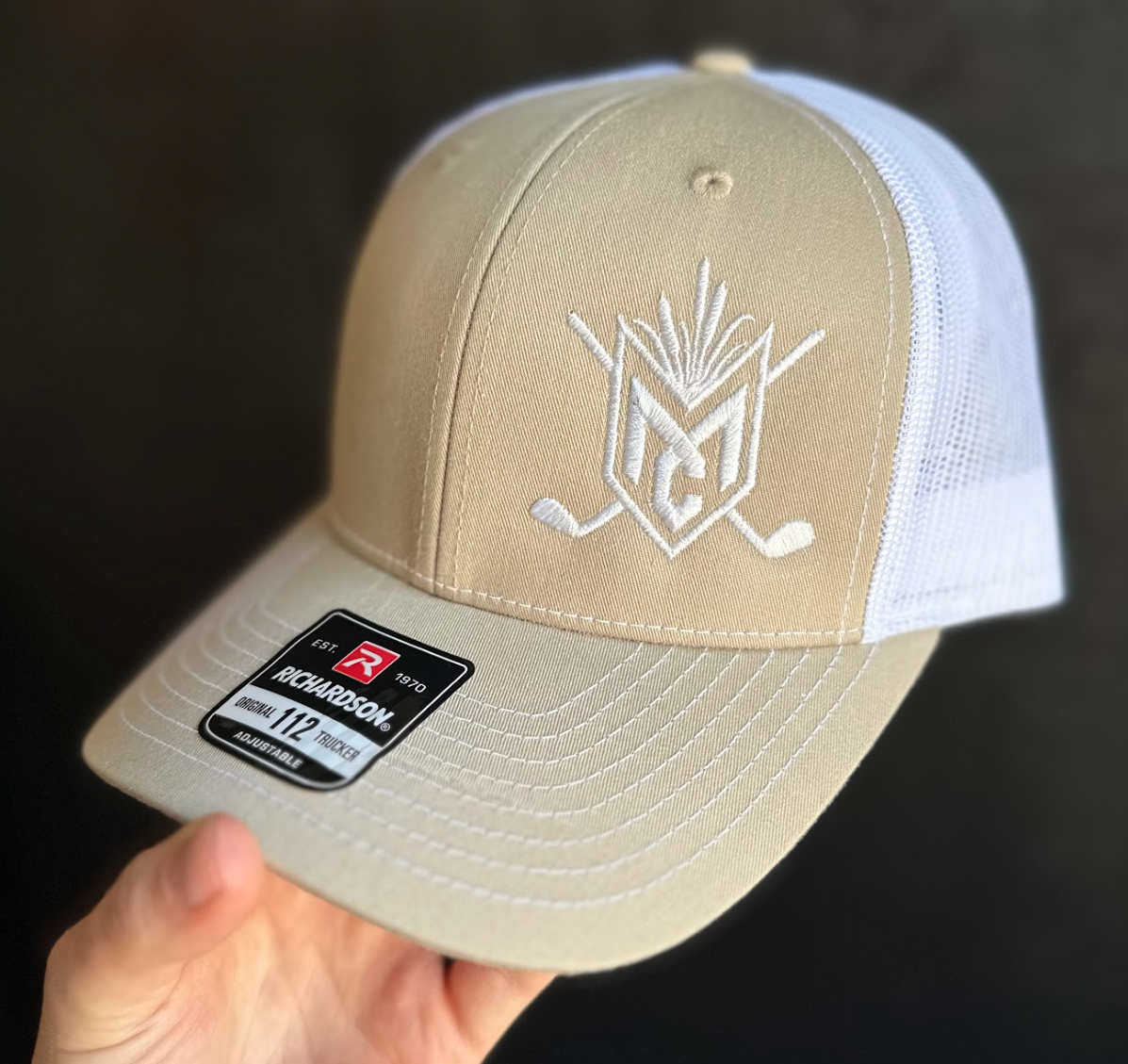 Beige and white cap with logo held by a hand against a dark background