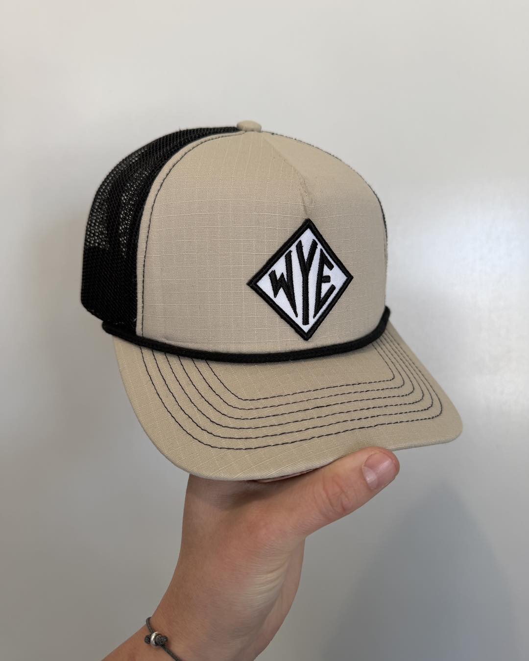 Beige and black cap with a logo held by a hand against a plain background WYE Angus Hat