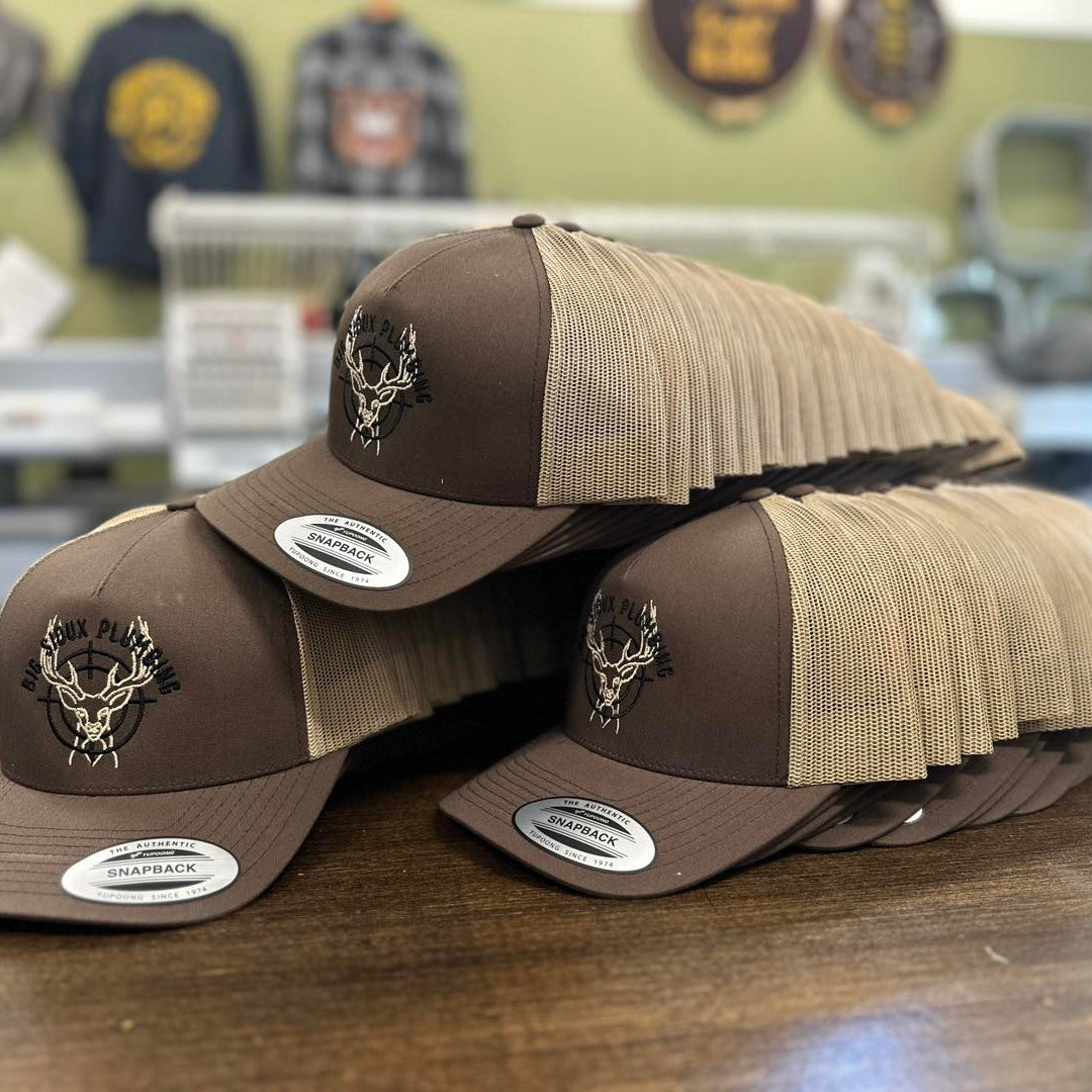 Stack of brown and beige YP Classic caps with deer logo on a wooden surface, Cotton & Custom's store setting in the background.