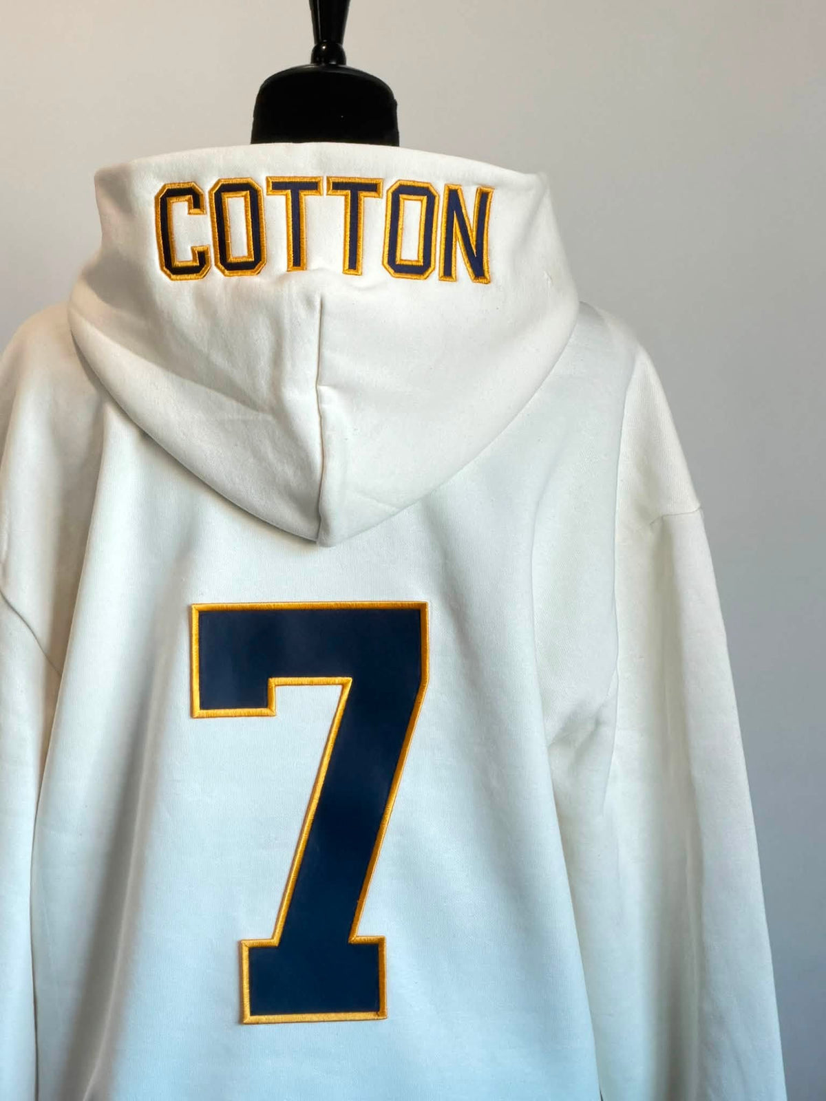 White hooded sweatshirt shown from the back on a mannequin, with 'COTTON' embroidered in navy and gold across the hood and a large navy number 7 outlined in gold on the back.