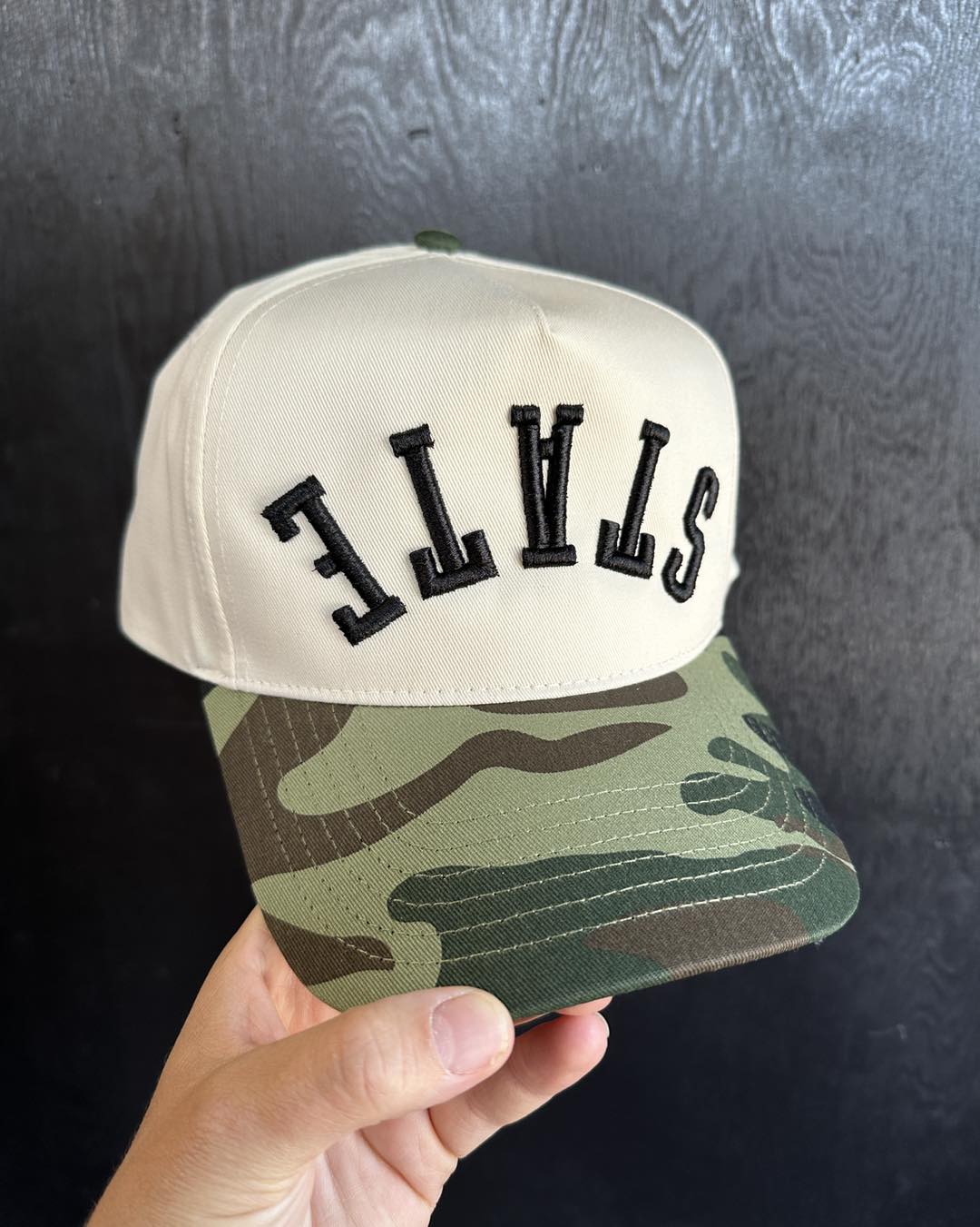 Cap with 'STATE' text on beige and camouflage design held against a dark background at cotton and customs llc.